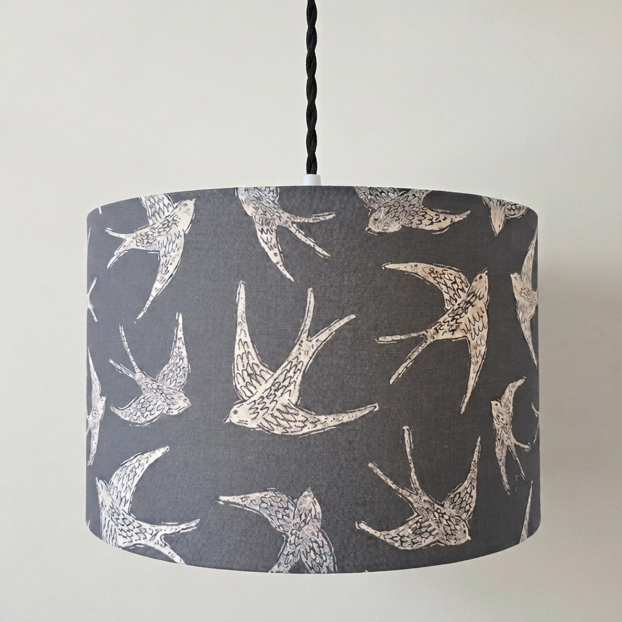A drum pendant lampshade with a dark gray background and a white printed pattern of flying swallows, suspended by a black cord from a white ceiling canopy against a plain off-white wall.