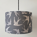 A drum pendant lampshade with a dark gray background and a white printed pattern of flying swallows, suspended by a black cord from a white ceiling canopy against a plain off-white wall.